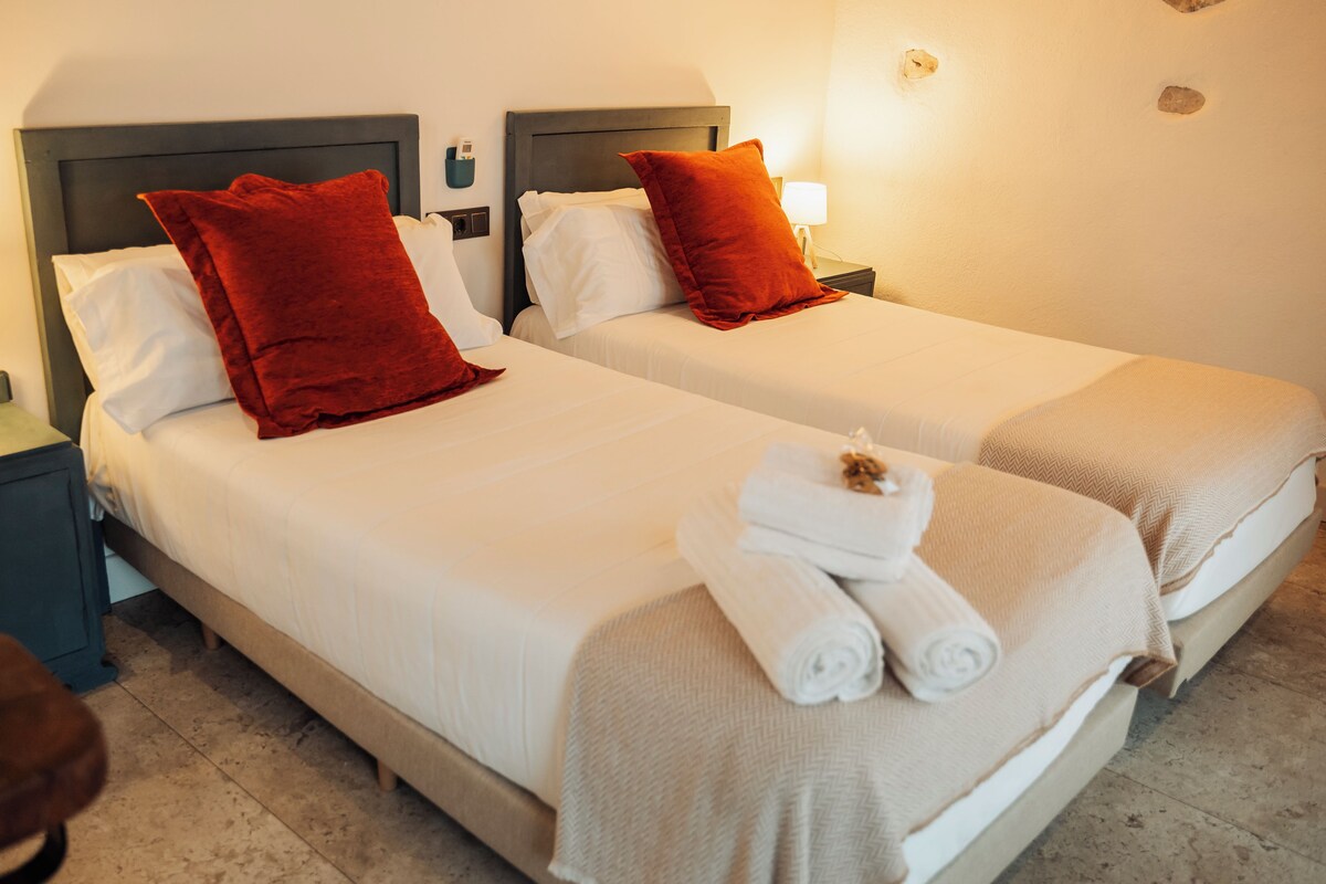A hotel room with two beds, each with white bedding and red pillows. There are two bedside tables with lamps and a small shelf with toiletries.