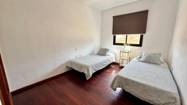 A room with two single beds, a small table, and a window with a blind.