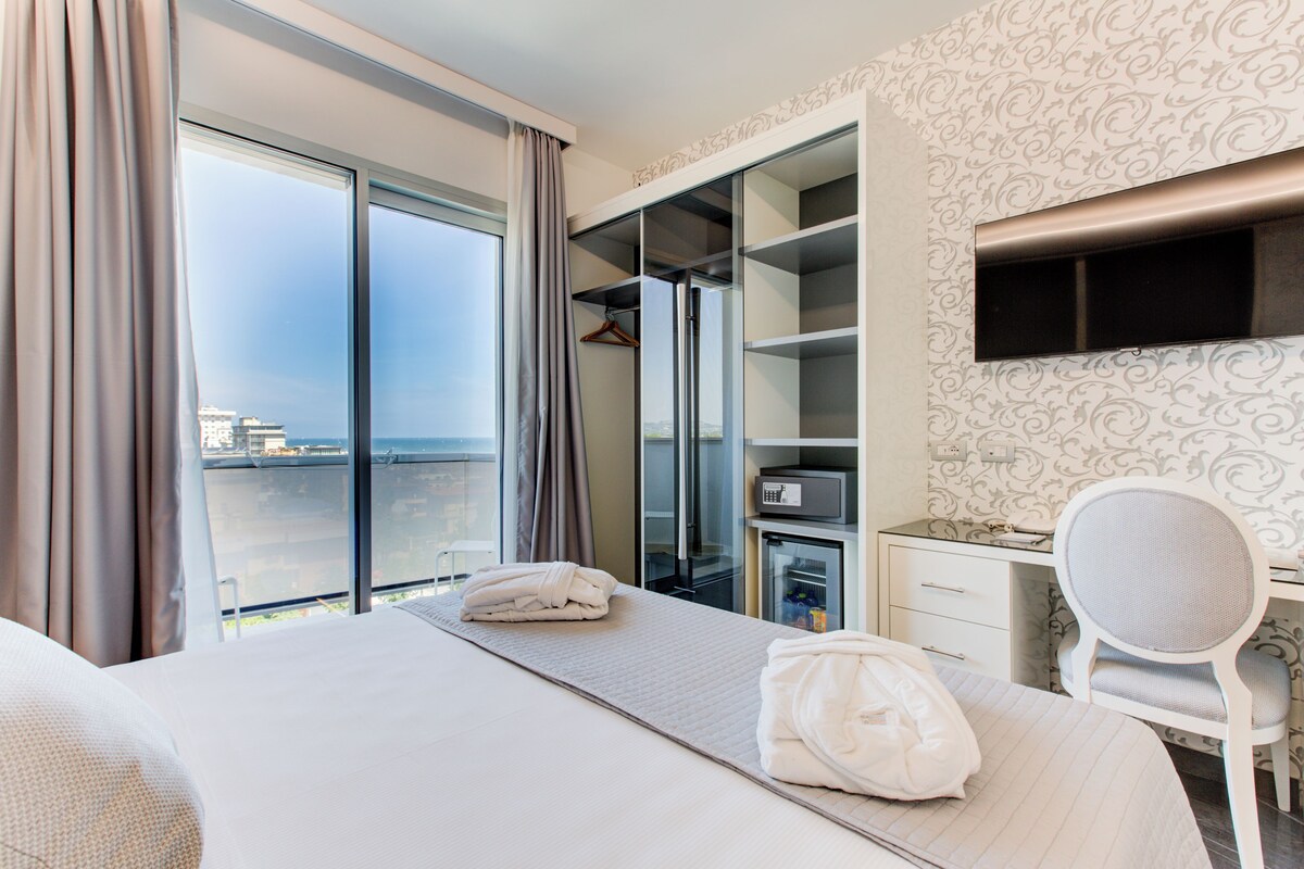 Habitación de hotel con cama, escritorio, silla, minibar y un ventanal con cortinas.