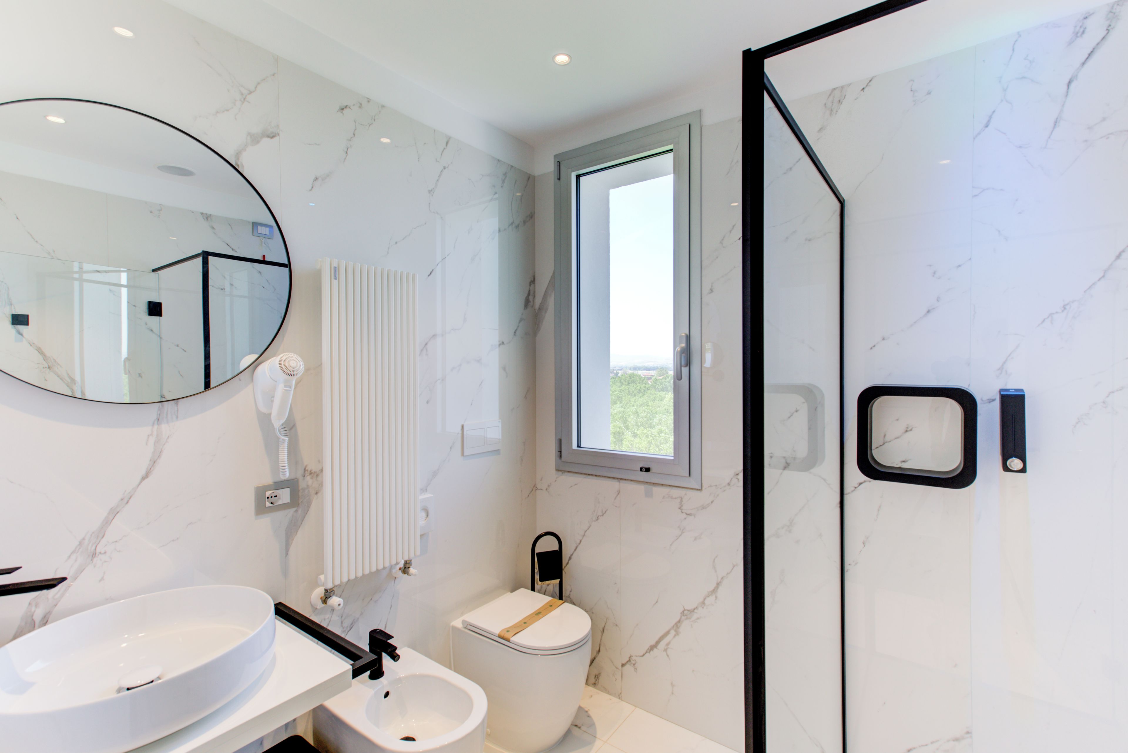 Un baño moderno con un espejo redondo, un lavabo blanco, un inodoro y una zona de ducha con mampara de vidrio.
