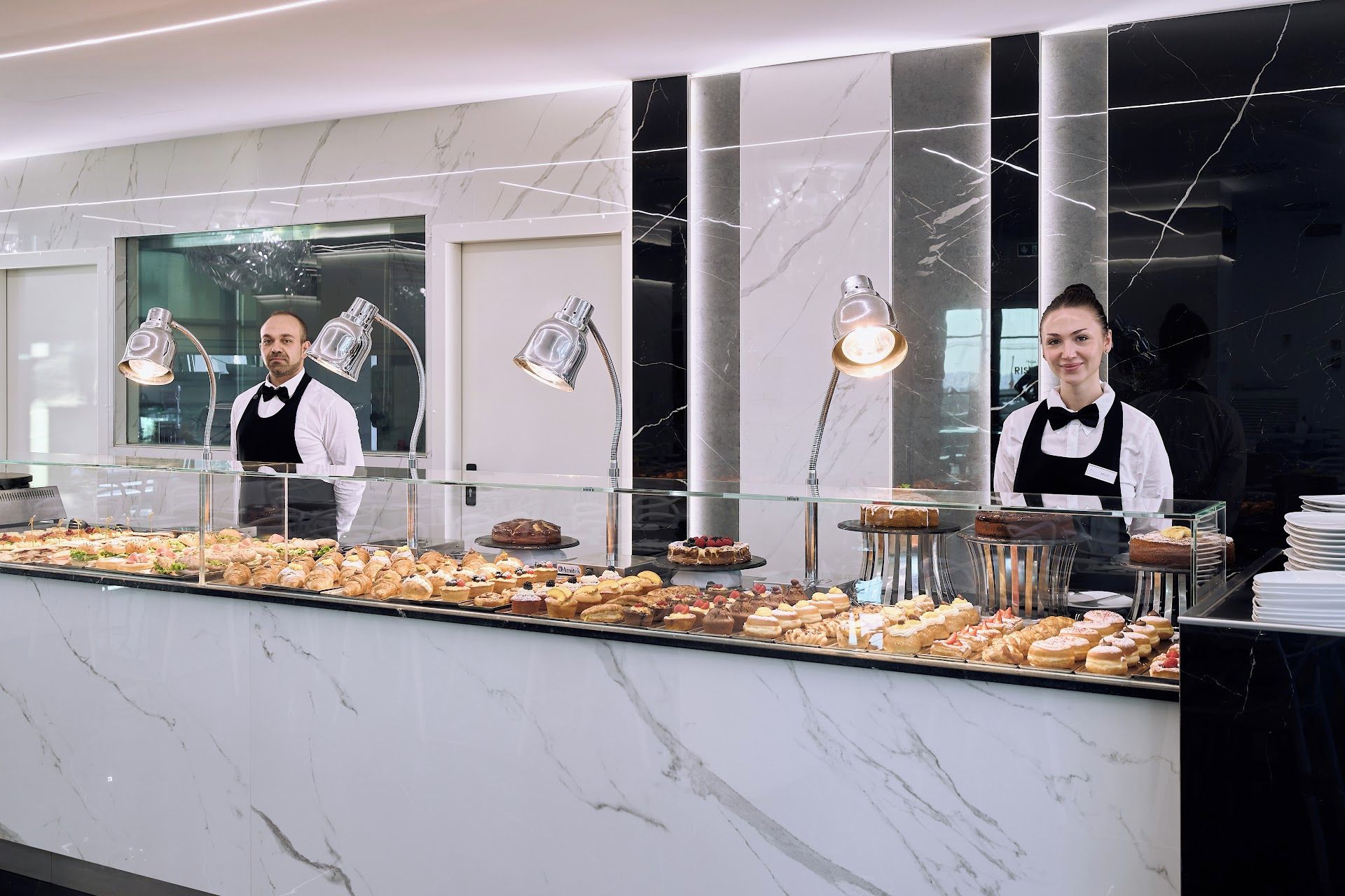 Mostrador de buffet con una variedad de pasteles y postres, una barrera de cristal reflectante y dos empleados detrás.