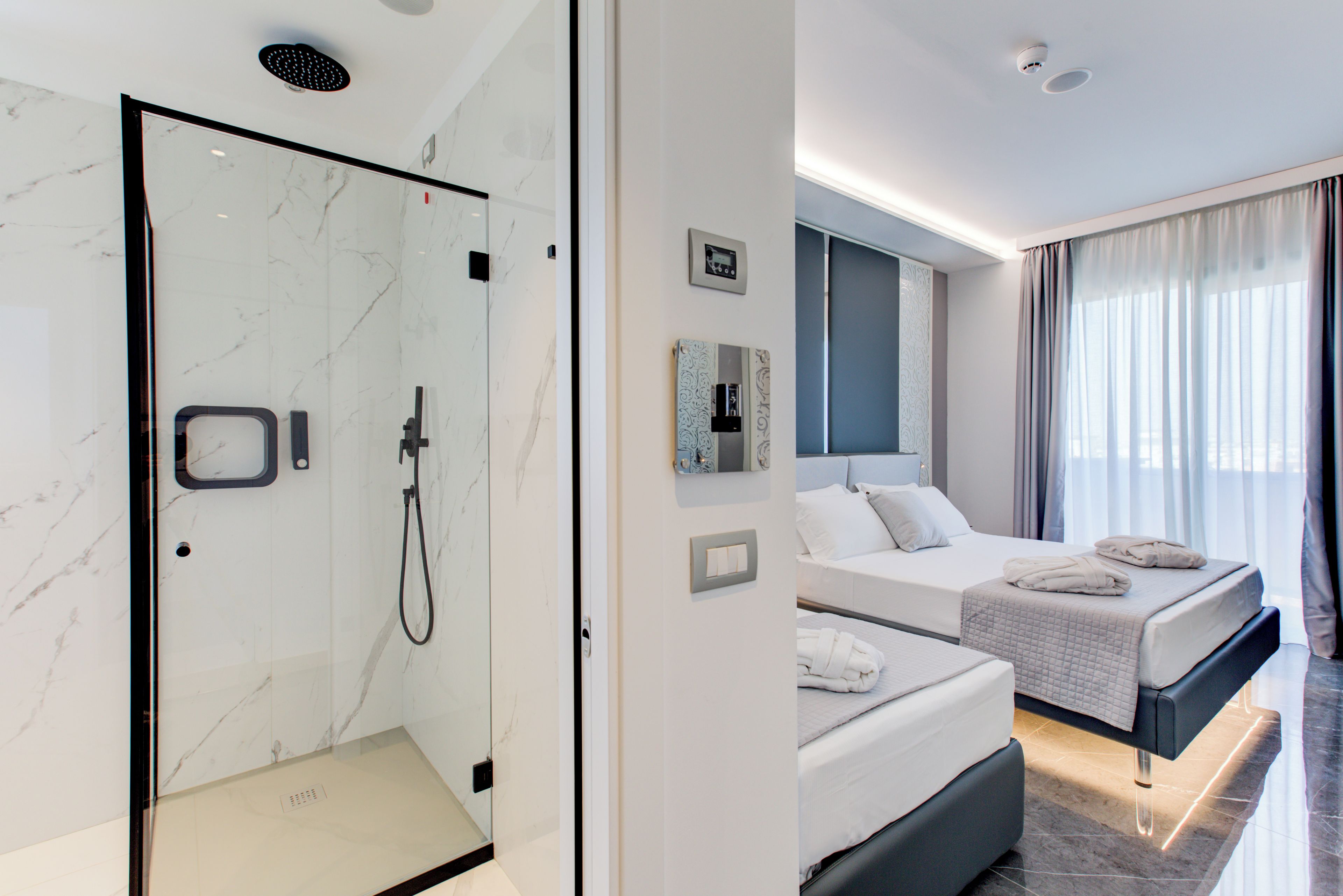 Una habitación de hotel moderna con ducha de vidrio, cama con cabecera gris y un ventanal con cortinas.