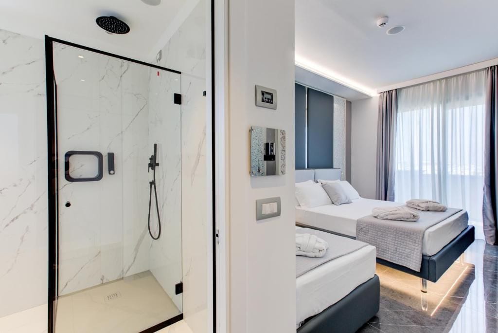 A modern hotel room with a glass-enclosed shower, a bed with a dark headboard, and a large window with curtains.