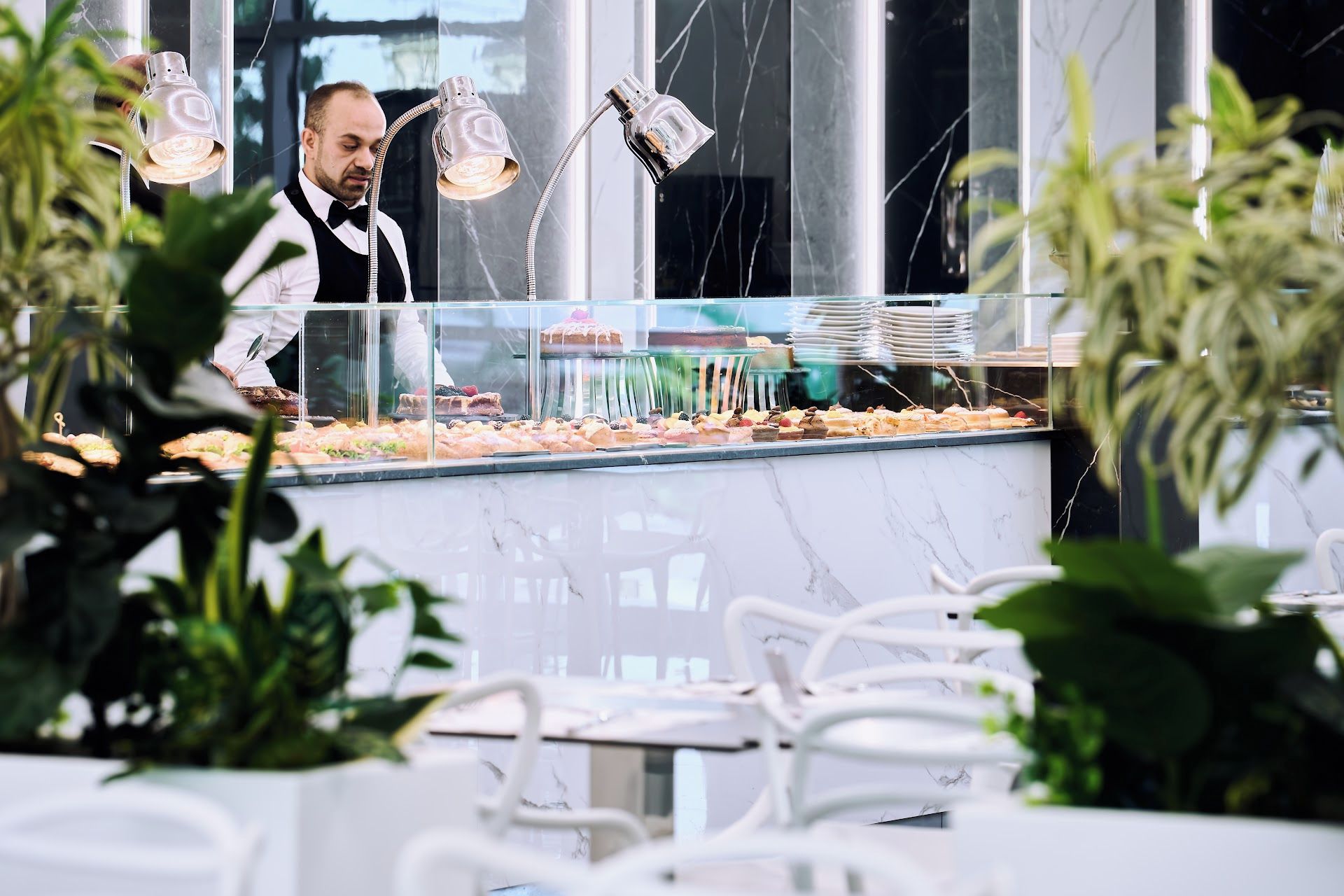 A restaurant with a marble counter displaying various desserts, a waiter in the background, and white chairs around the dining area.