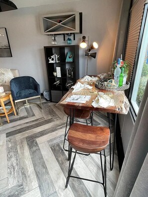 A dining area with a wooden table, two chairs, a blue armchair, and a black bookshelf. - Couples Getaway 105 -1BR Gold Coast, Cherry Grove (North Myrtle Beach)