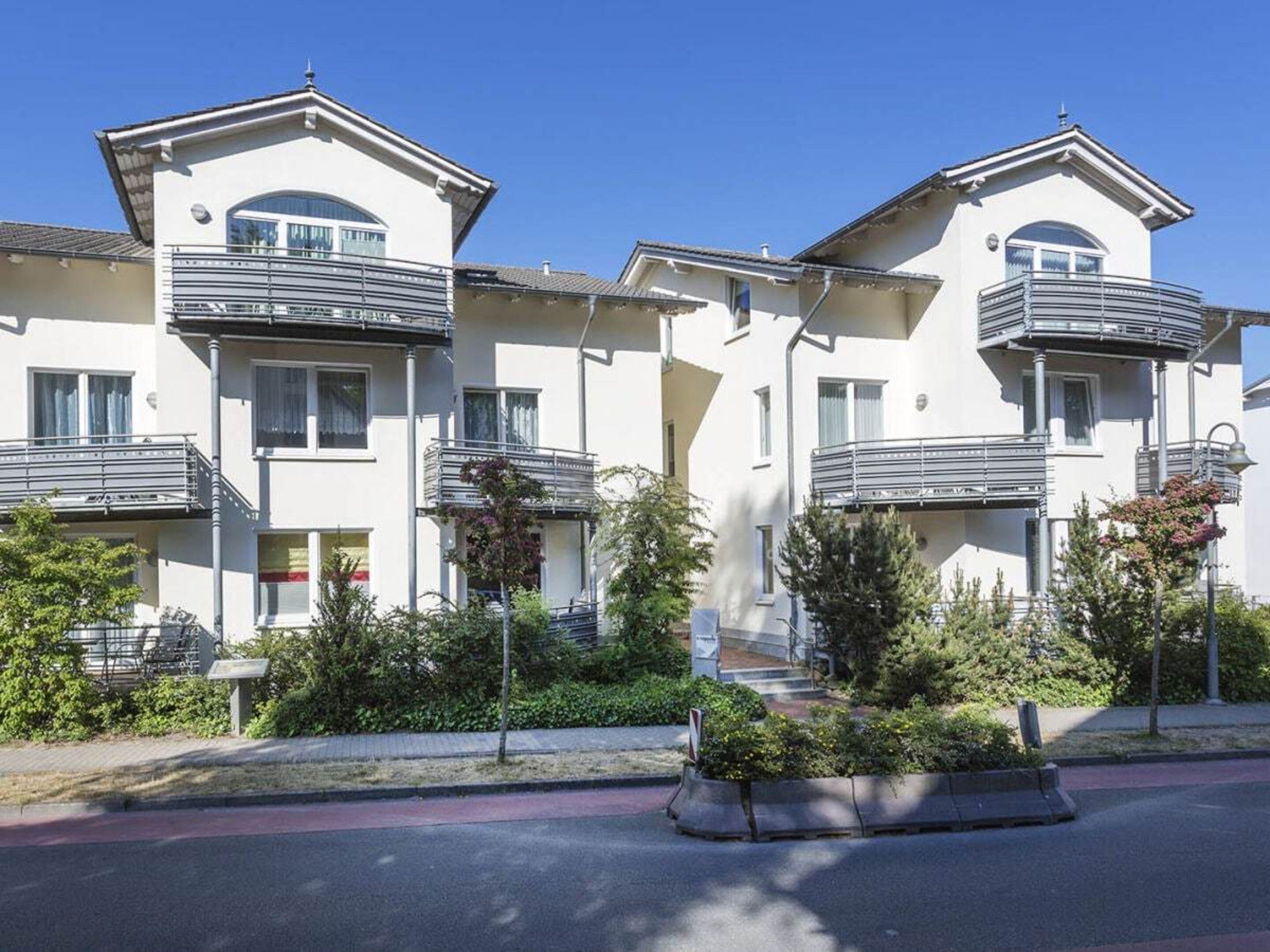 A row of modern multi-story residential buildings with balconies, a landscaped area with shrubs, and a clear sky.