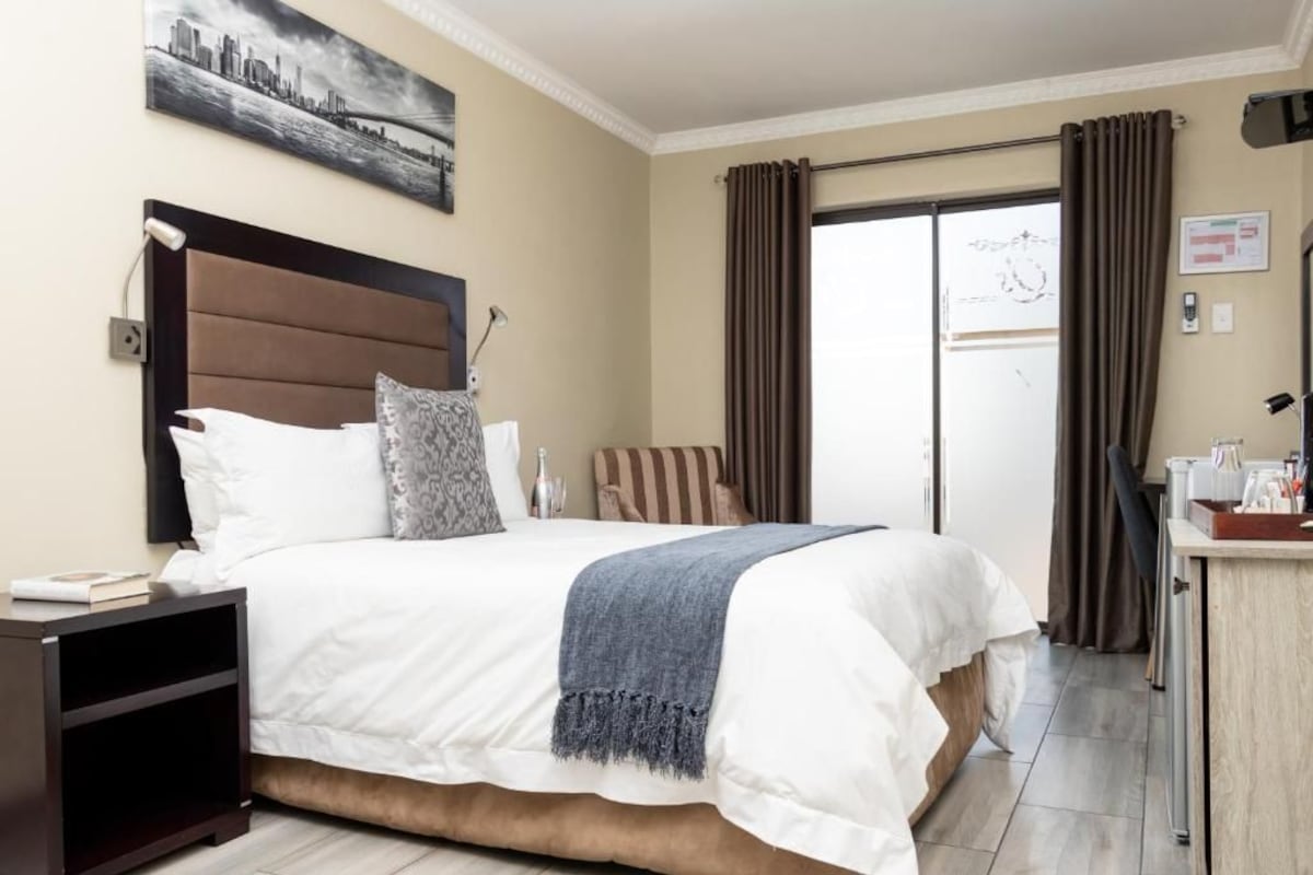 Habitación de hotel con una cama grande, una mesita de noche, un escritorio y vistas al exterior a través de una puerta de vidrio.