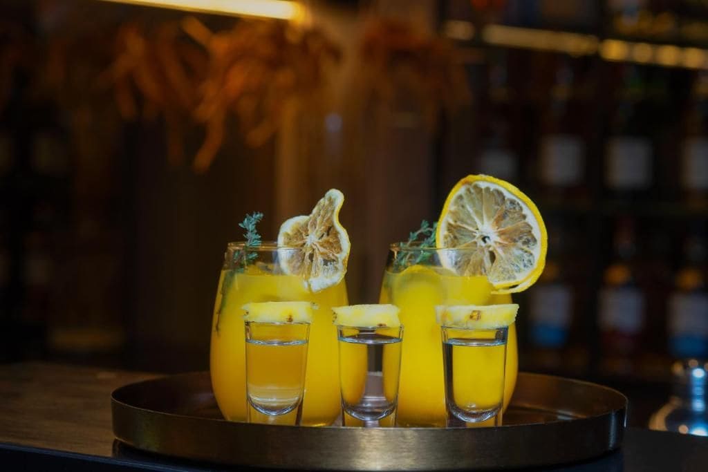 Un vassoio con tre bicchieri contenenti una bevanda gialla, ognuno guarnito con una fetta di limone e un rametto di erbe aromatiche.