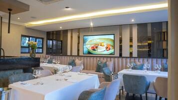 Un restaurante con varias mesas, asientos acolchados y una pantalla grande que muestra un plato.