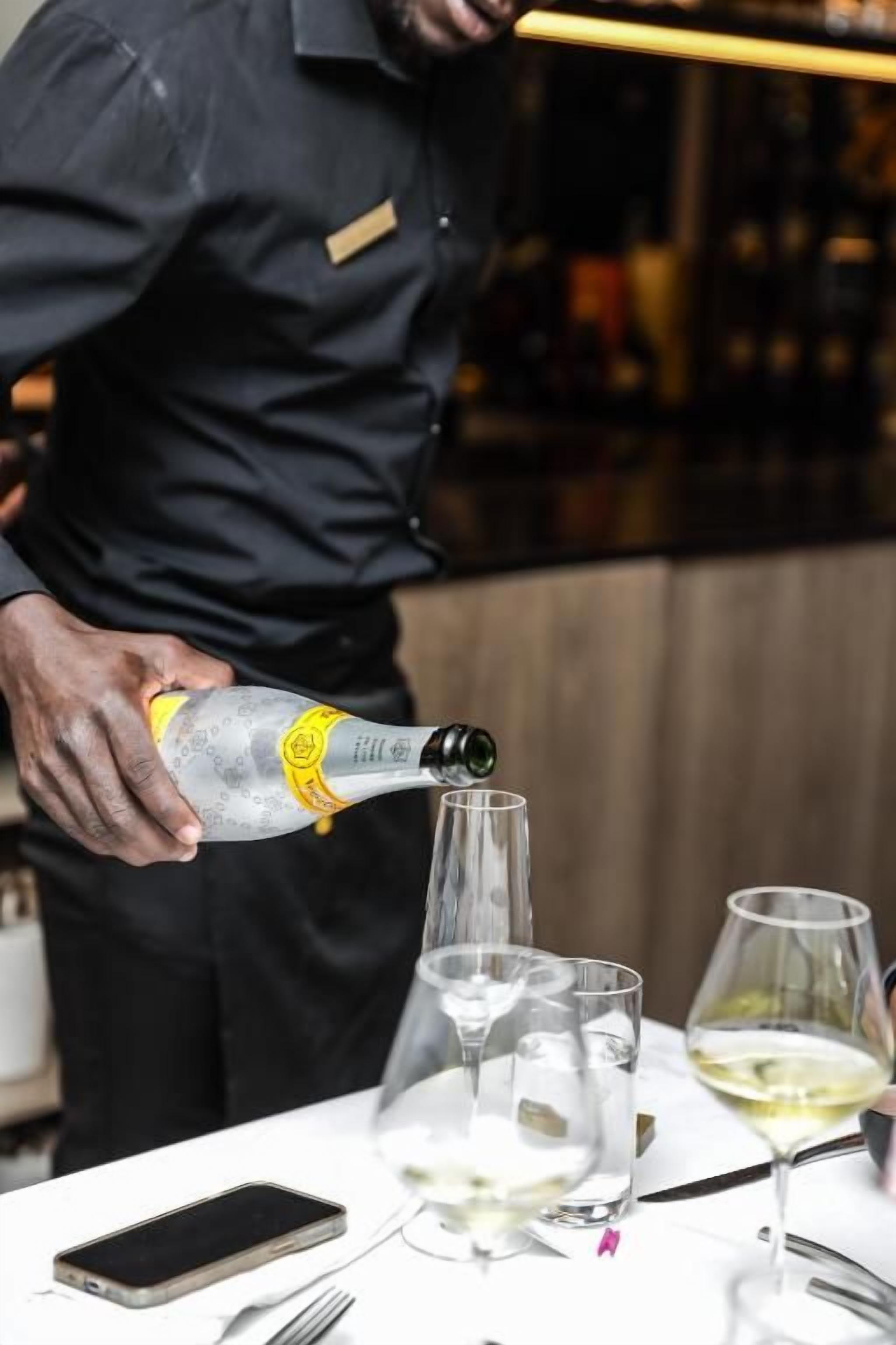 A person pouring a drink into a glass on a table with a smartphone, fork, and knife.
