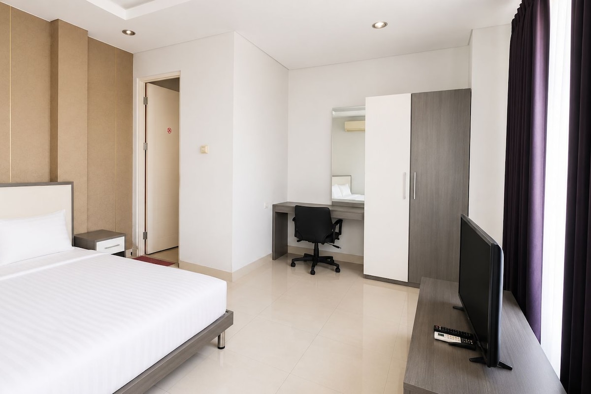 A hotel room with a large bed, a desk with a chair, a television, and a bathroom visible through an open door.