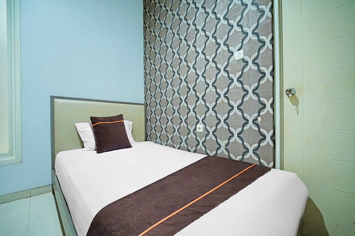 Een enkel bed met een witte matras, een bruine deken en twee kussens in een kamer met lichtblauwe muren en behang met een patroon.