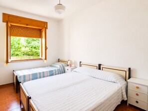 Ein Schlafzimmer mit zwei Betten, einem Fenster mit Holzjalousie, einem Nachttisch mit Lampe und einem zusätzlichen Nachtschränkchen.