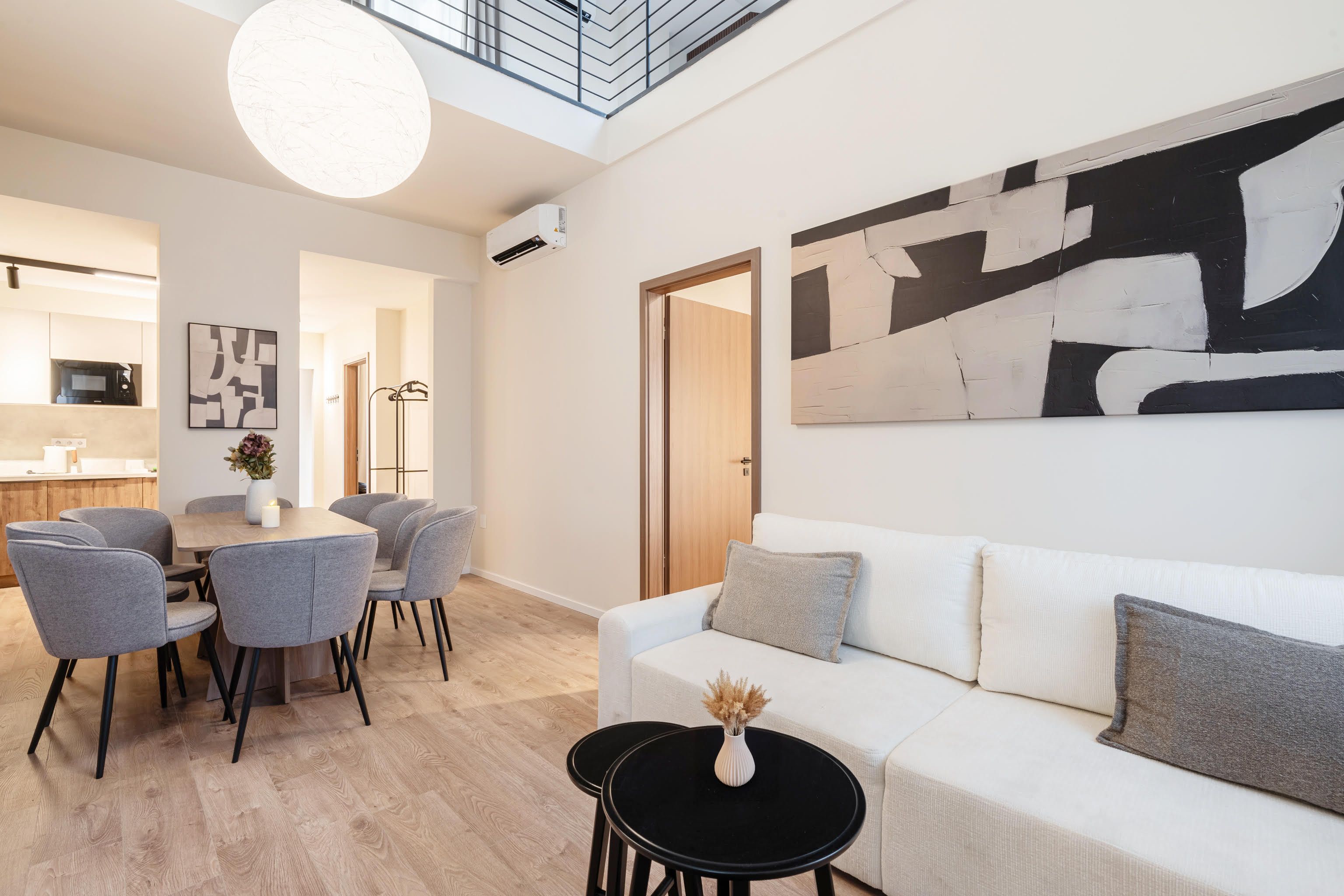 Un salón moderno con un sofá blanco, sillones grises y una mesa de centro negra. Hay un cuadro abstracto grande en la pared, una zona de comedor con mesa y sillas, y una cocina al fondo.