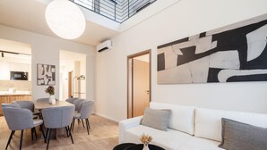 A modern living room with a white sofa, grey armchairs, and a black coffee table. There is a large abstract painting on the wall, a dining area with a table and chairs, and a kitchen in the background. - LimeTree Residence by BQA (Budapest)