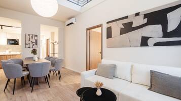 Un salón moderno con un sofá blanco, sillones grises y una mesa de centro negra. Hay un cuadro abstracto grande en la pared, una zona de comedor con mesa y sillas, y una cocina al fondo.