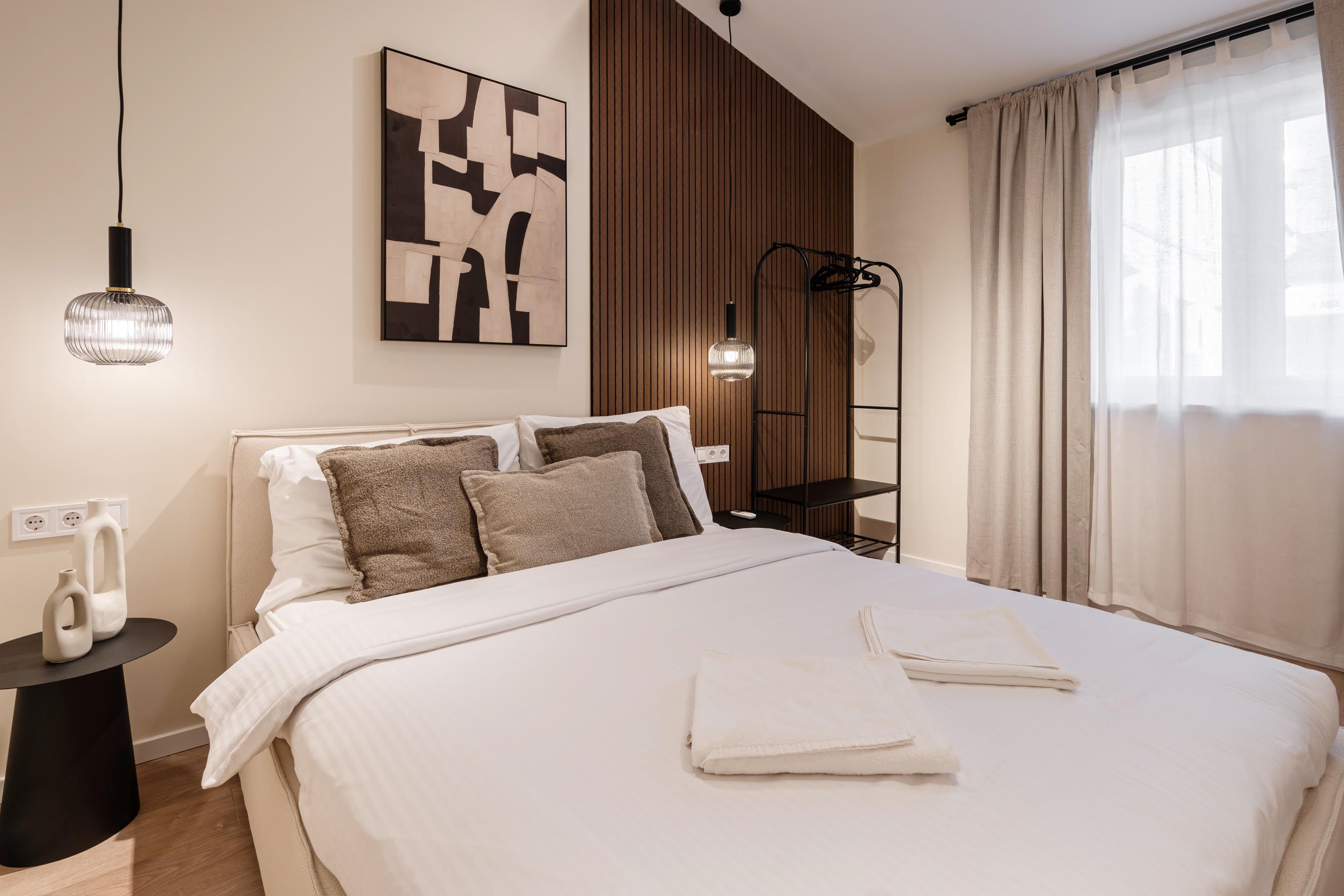 Una cama bien hecha con sábanas blancas y dos toallas, un cuadro abstracto moderno en la pared, una lámpara de pie y un panel de pared de madera.