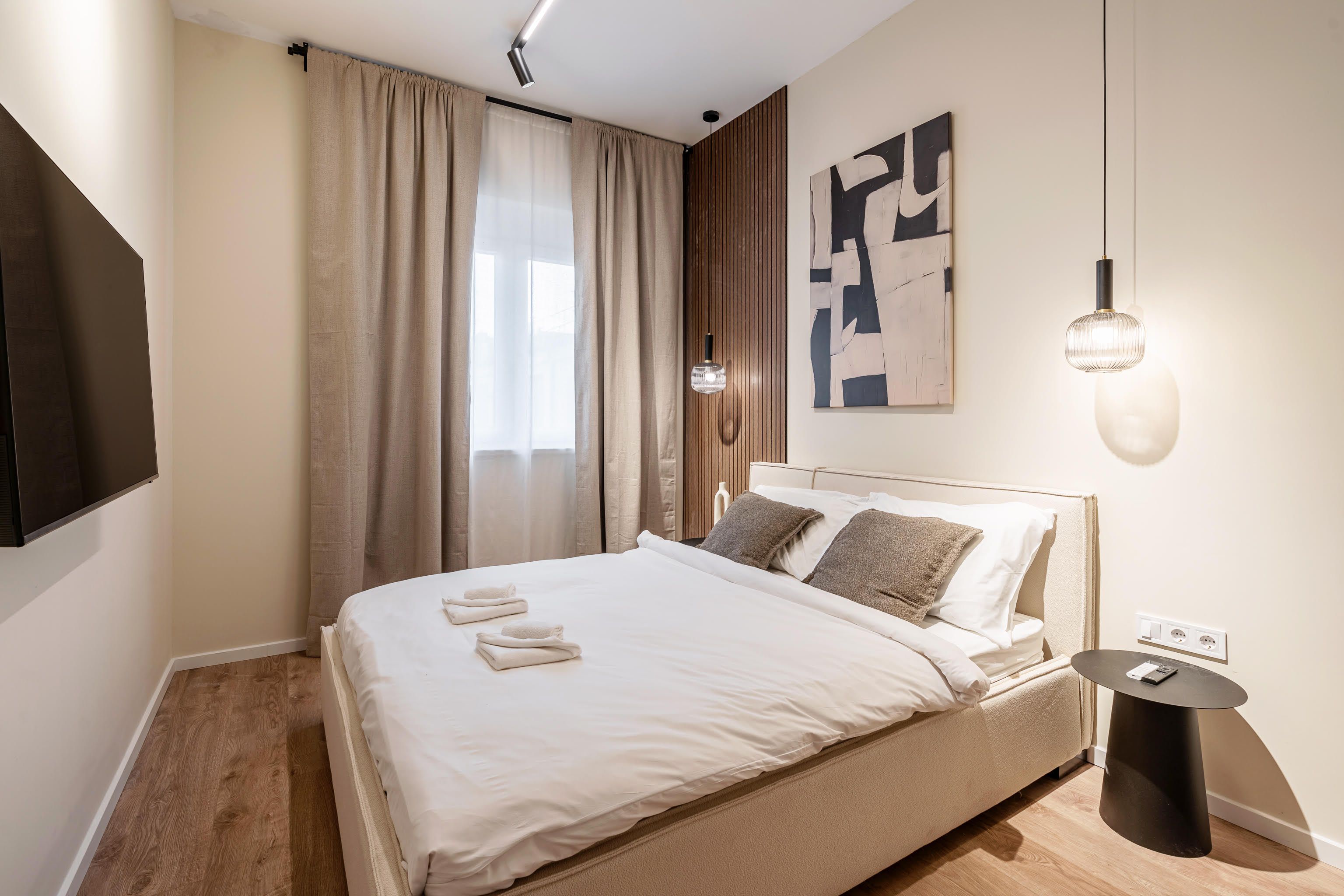 A modern hotel room with a large bed, a flat-screen TV, a bedside table, and a wall-mounted light fixture.