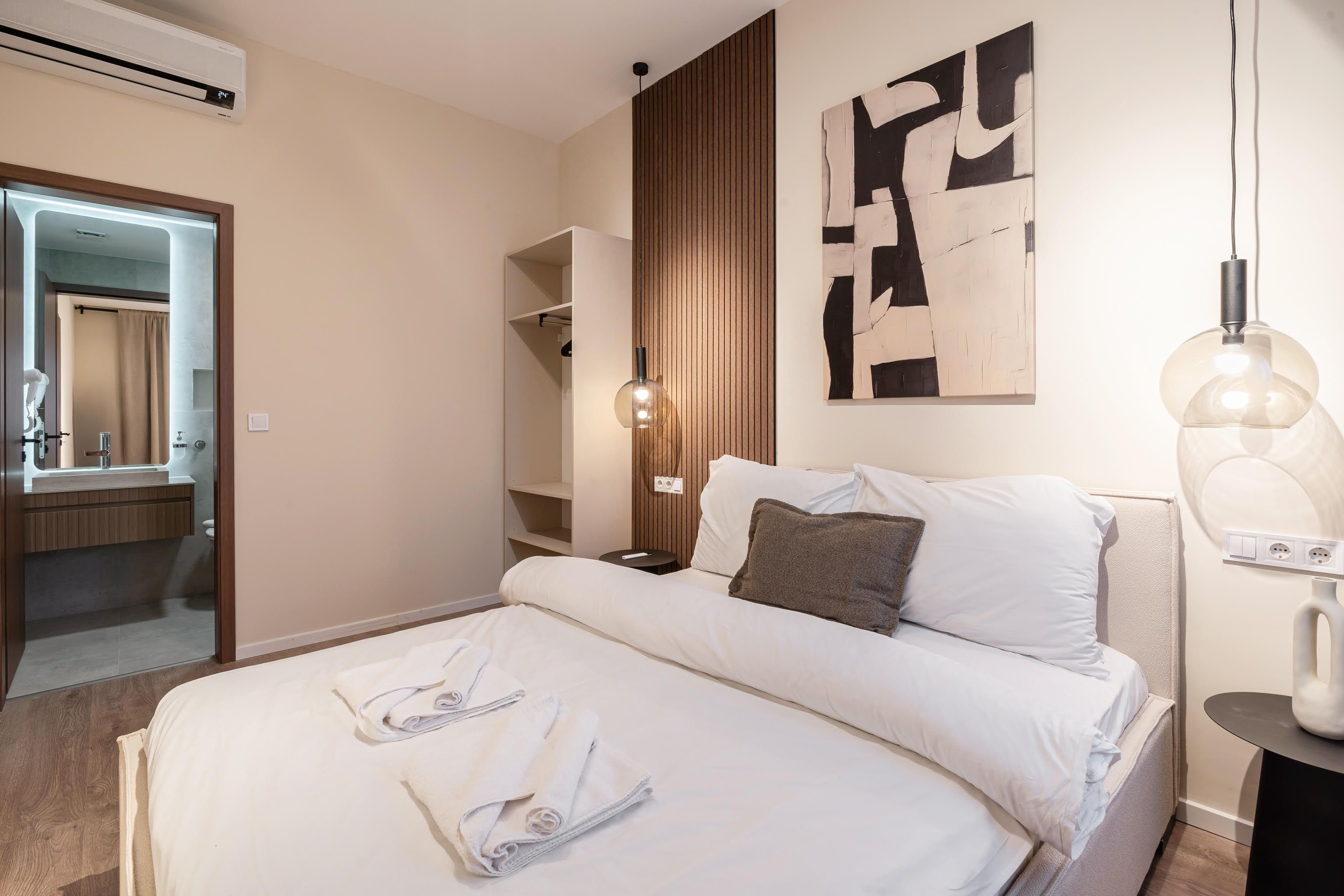 Habitación de hotel con cama, mesita de noche, un cuadro en la pared, lámpara colgante y un baño visible a través de una puerta abierta.
