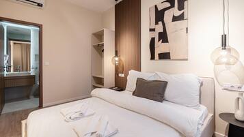 Habitación de hotel con cama, mesita de noche, un cuadro en la pared, lámpara colgante y un baño visible a través de una puerta abierta.