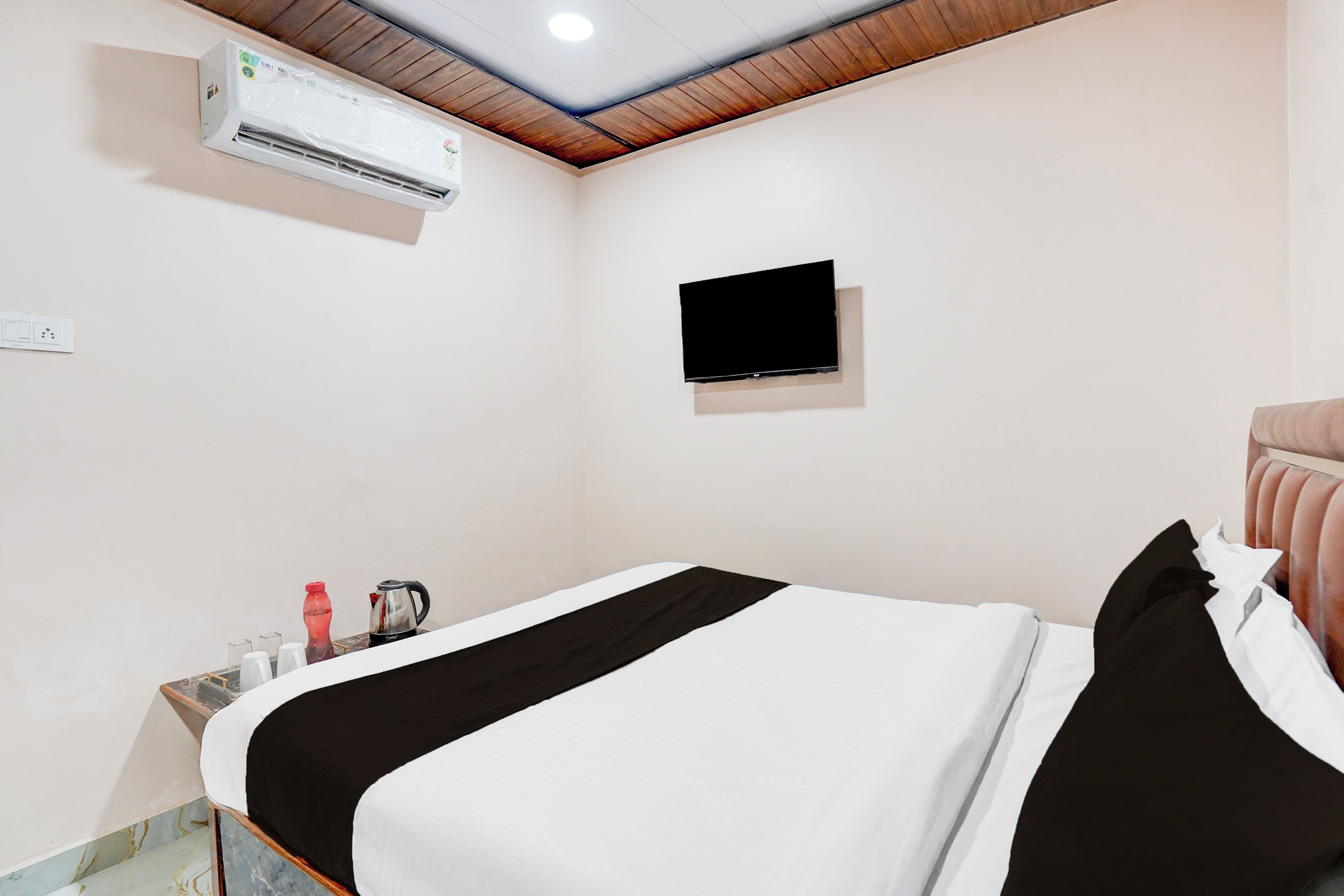 Habitación de hotel con cama, televisor montado en la pared, aire acondicionado y una pequeña mesa con tetera y tazas.