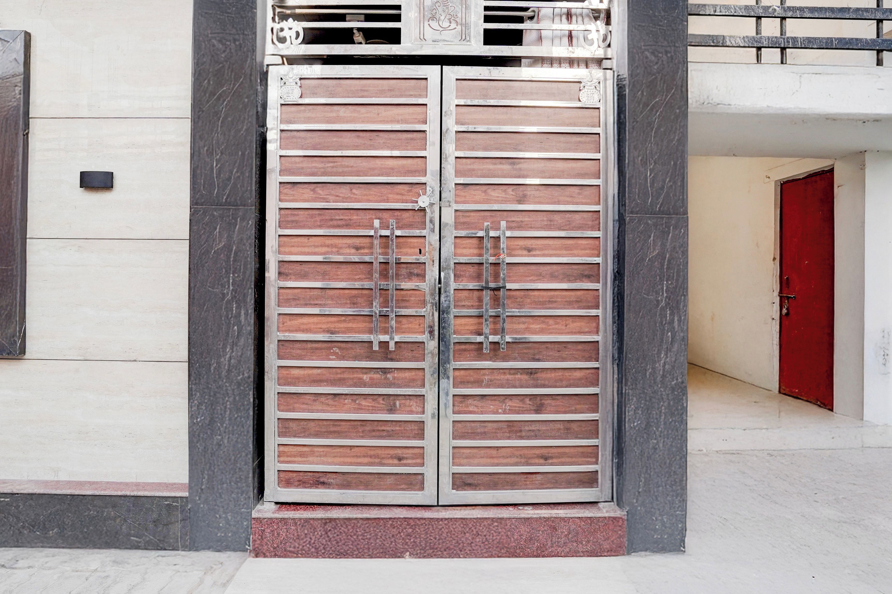 Una reja doble de metal con barras horizontales y diseño de paneles de madera, frente a una fachada de edificio donde se ve una puerta roja al fondo.