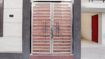 Una reja doble de metal con barras horizontales y diseño de paneles de madera, frente a una fachada de edificio donde se ve una puerta roja al fondo.