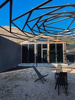 A modern building with a metal framework, large glass windows, and a gravel patio with outdoor furniture.