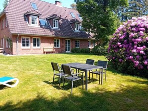 A house with a sloped roof, a lawn, a table with chairs, and a flowering bush. - Holiday Apartment in Nebel With Swimming Pool (Nebel)