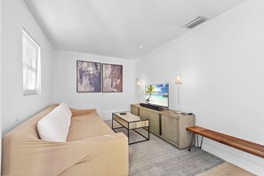 A modern living room with a beige sofa, a coffee table, a TV on a wooden stand, and artwork on the wall. - Tropical 1BR Casita w/ Shared Pool Access (West Palm Beach)