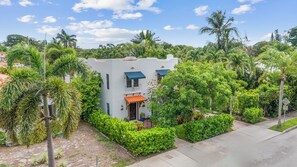 A two-story building with a white facade and blue awnings, surrounded by lush greenery and palm trees. - Tropical 1BR Casita w/ Shared Pool Access (West Palm Beach)