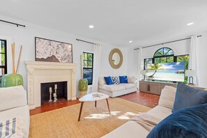 A modern living room with a fireplace, a large flat-screen TV, and a view of a beach through the window. - 4BR Pool Home w/ Private Guest Suite – West Palm Beach (West Palm Beach)