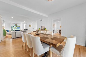 A spacious dining area with a wooden table and white chairs, a kitchen in the background, and a living area with a sofa and a TV. - Tropical 3BR Oasis w/ Pool (West Palm Beach)