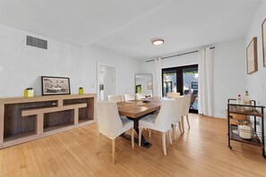 A modern dining area with a wooden table and white chairs, a whiteboard, and a small shelf with items. - Tropical 3BR Oasis w/ Pool (West Palm Beach)
