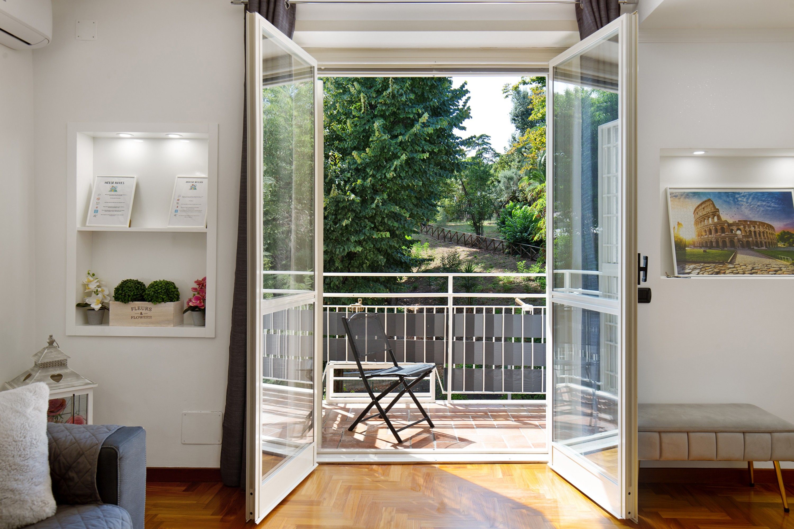 An open balcony with a chair, a wooden floor, and a view of a garden with trees and a fence.
