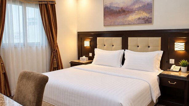 A hotel room with a large bed, a bedside table with a lamp, a chair, and a painting on the wall. - Manazilna Serviced Apartments (Riyadh)
