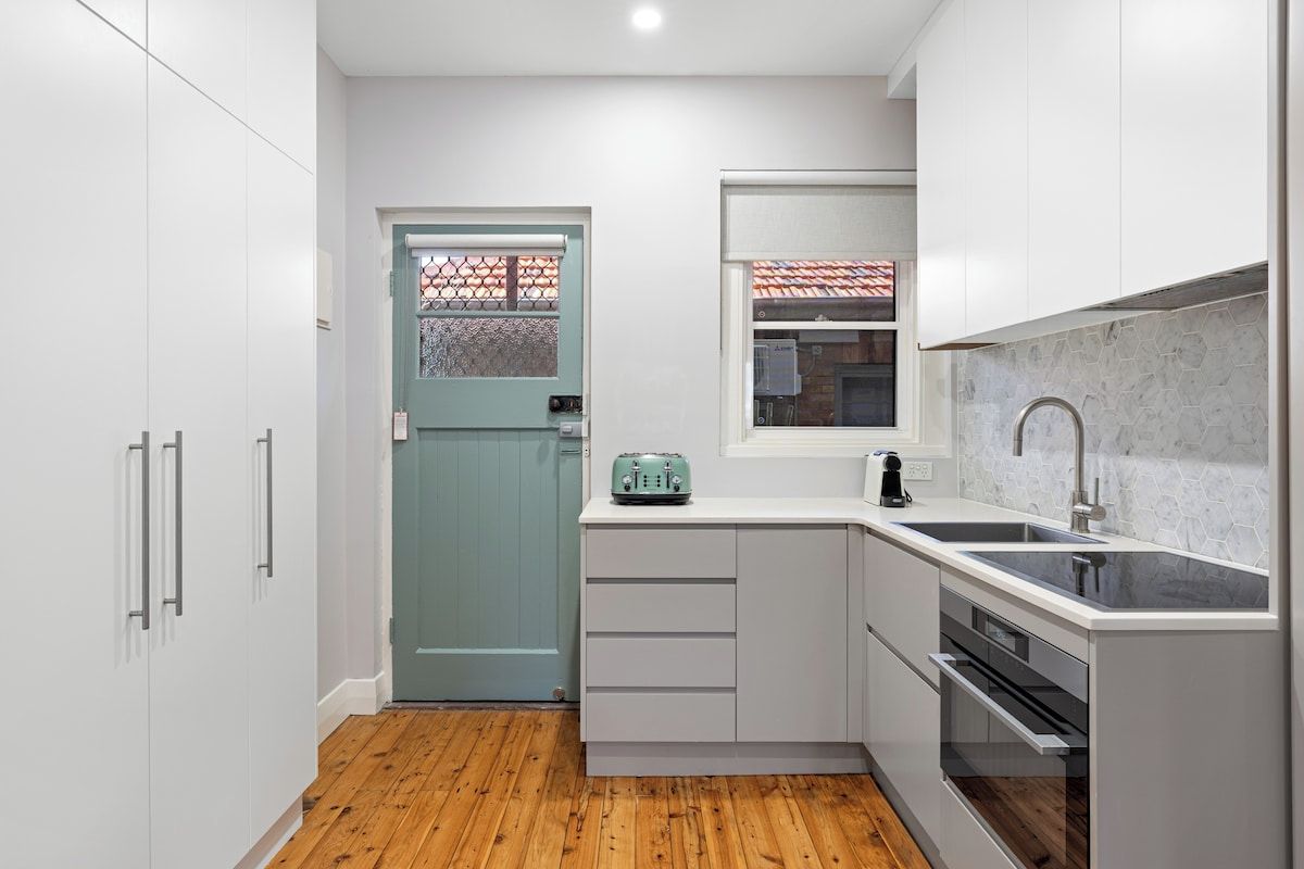 A compact kitchen with white cabinetry, a wooden floor, a teal door, and a window with a tiled roof view.