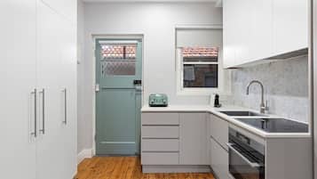 A compact kitchen with white cabinetry, a wooden floor, a teal door, and a window with a tiled roof view.