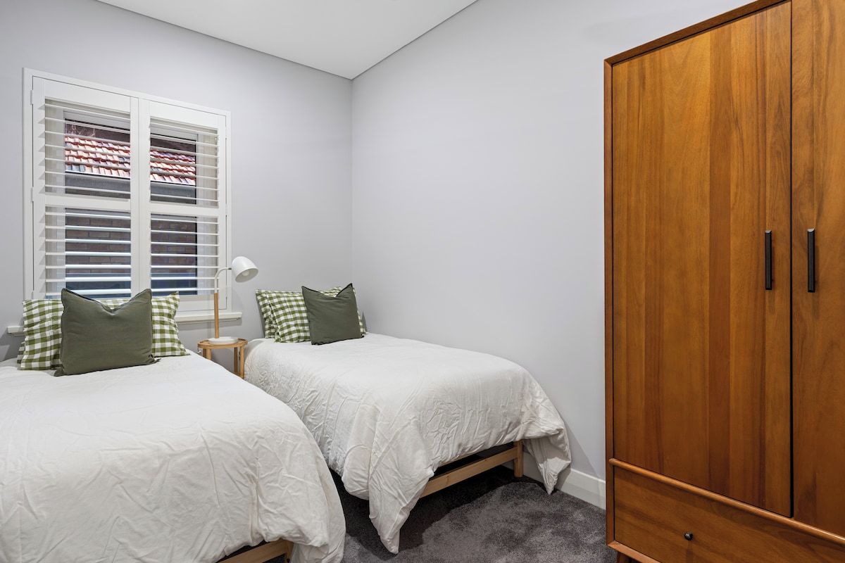 A bedroom with two single beds, a wardrobe, a window with shutters, and a bedside table with a lamp.