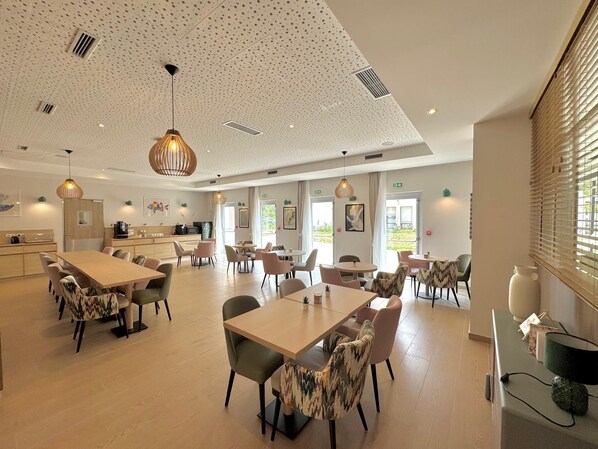 A spacious dining area with multiple tables and chairs, a kitchen in the background, and a ceiling with a perforated pattern.