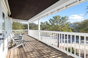 A covered wooden deck with white railings, outdoor furniture, and a view of trees and a building in the background.