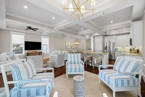 A spacious living area with a large television, white sofas, striped armchairs, and a kitchen area with modern appliances.