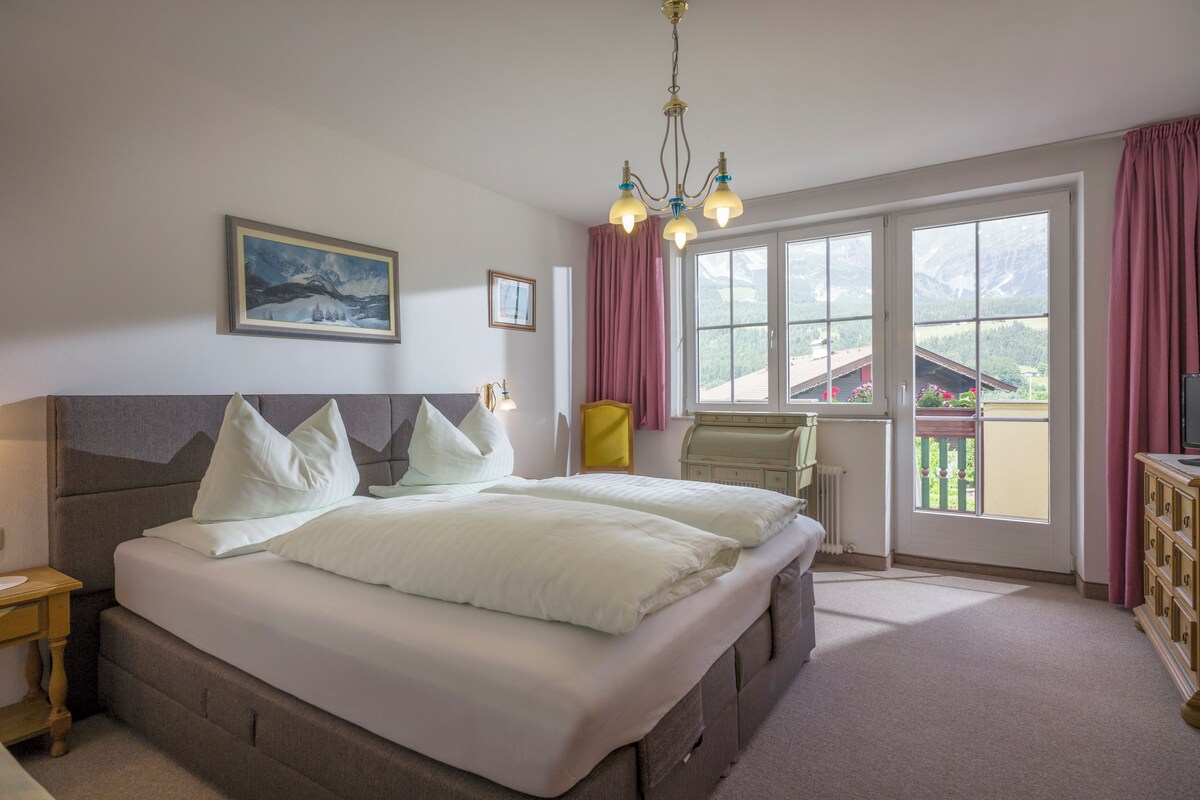 A bedroom with a large bed, a headboard, a bedside table, a chair, a window with curtains, and a view of the outside.
