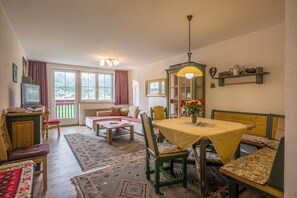 A cozy living room with a sofa, a dining table with chairs, a television, and a window with curtains. - Apparthotel Tom Sojer (Tirol)