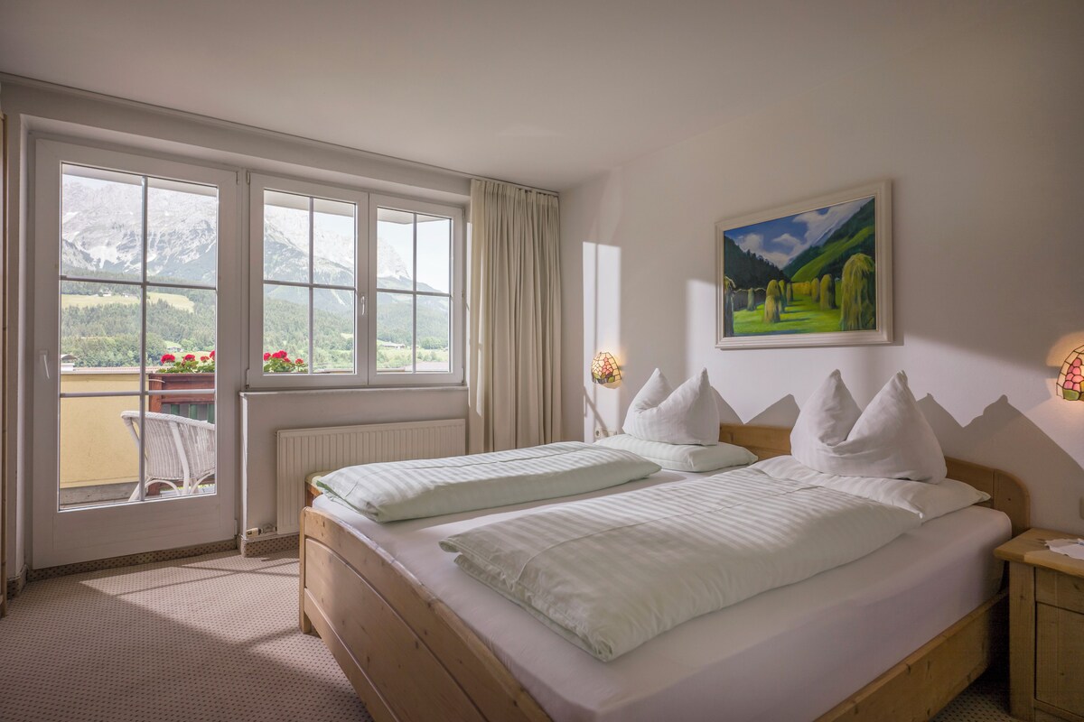 A bedroom with a large bed, a view of mountains through the window, and a painting on the wall.