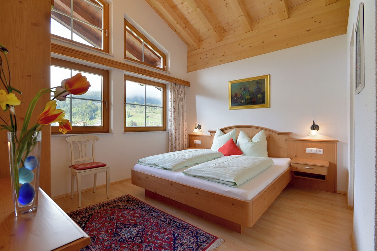 A bedroom with a wooden bed, a nightstand, a chair, a window with a view of the outdoors, and a painting on the wall.