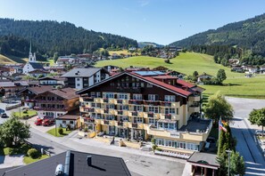 A multi-story hotel with a red roof, balconies with flower boxes, and a view of a town and mountains in the background.