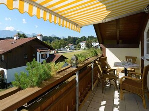 A balcony with a view of houses and mountains, wooden furniture, and a yellow-striped awning. - 3 - Ellen Raith (Fuessen)