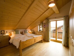 A bedroom with a wooden ceiling, a bed, bedside tables, a window with curtains, and a view of a wooden building outside. - 3 - Ellen Raith (Fuessen)