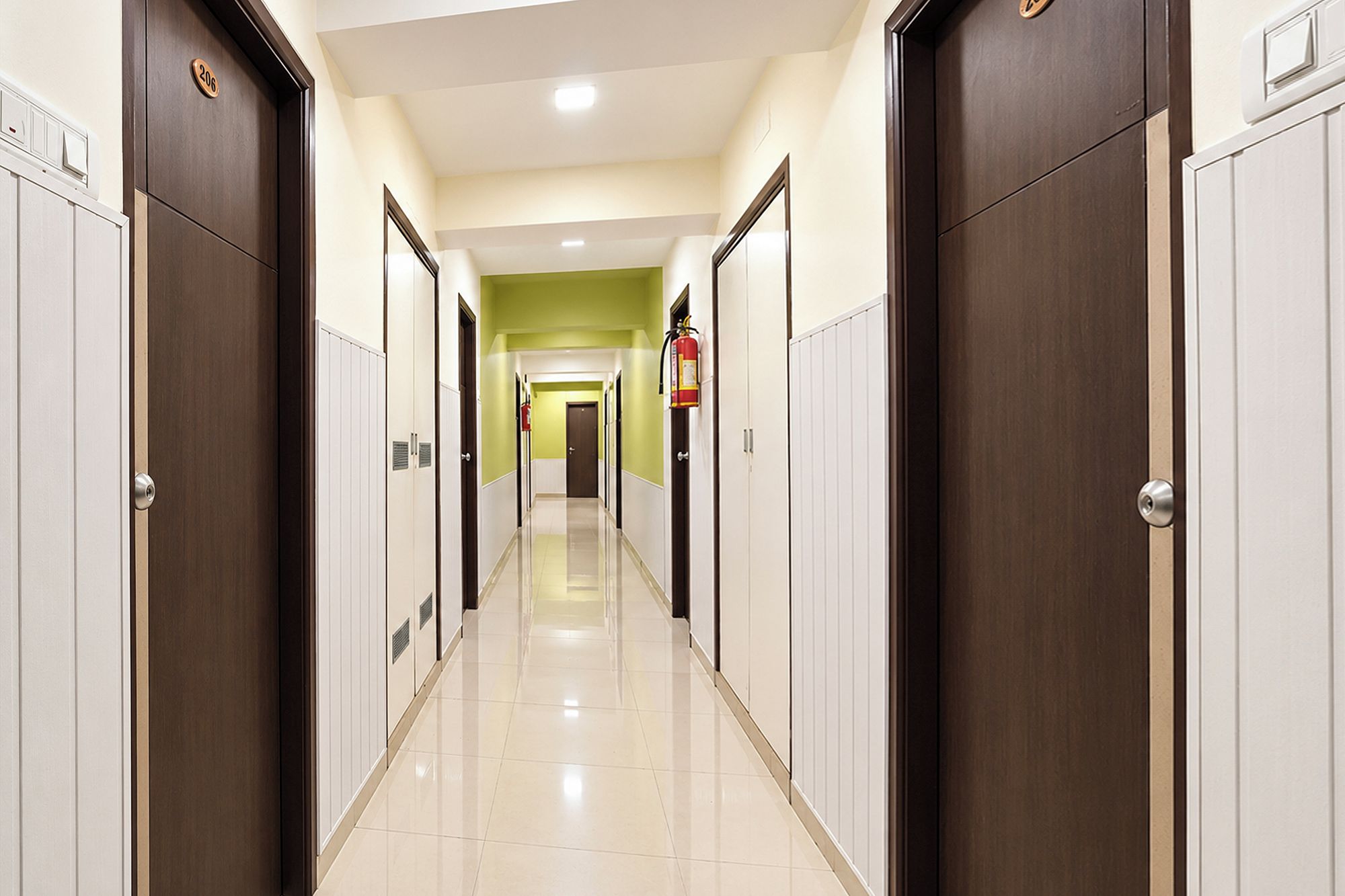 Corredor de hotel com portas em ambos os lados, um extintor de incêndio fixado na parede e piso revestido de azulejos.